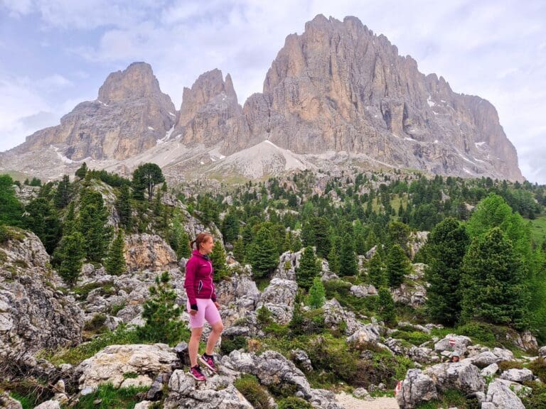 A person in a pink and purple outfit stands on rocky terrain with pine trees scattered around. Towering cliffs and a cloudy sky form the backdrop, creating a dramatic and scenic mountain landscape.
