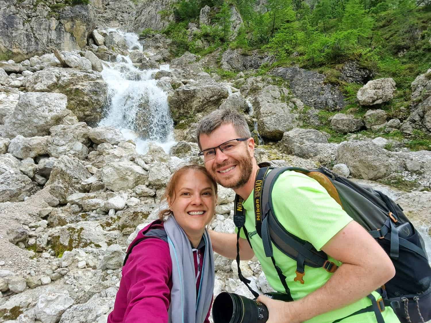 A couple takes a selfie near a rocky waterfall. The man wears a light green shirt and a backpack, holding a camera. The woman is in a pink jacket. Lush greenery is visible in the background. They both smile at the camera.