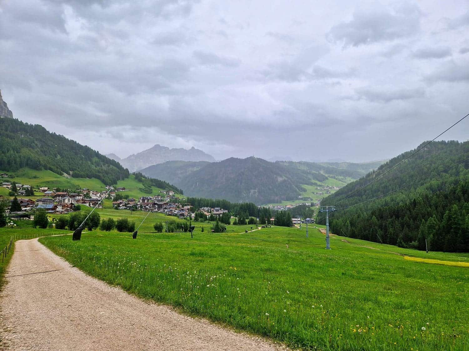 A scenic view of a small village nestled in lush green hills under a cloudy sky. A dirt path winds through grassy fields on the left, and there are distant mountains partially obscured by clouds.