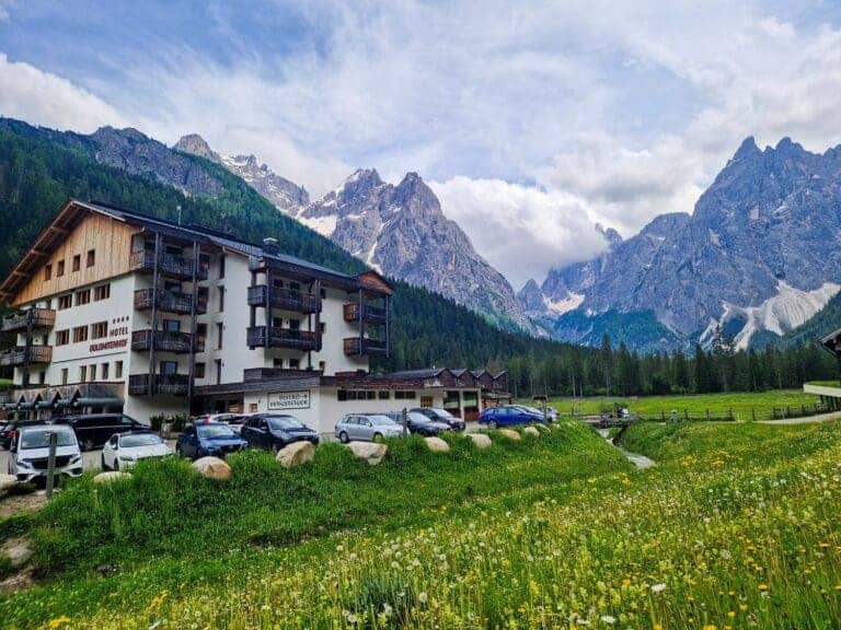 Mountain hotel in a scenic alpine valley surrounded by lush greenery and wildflowers. Cars are parked outside the multi-story wooden building. Majestic, cloud-capped mountains rise in the background under a partly cloudy sky.