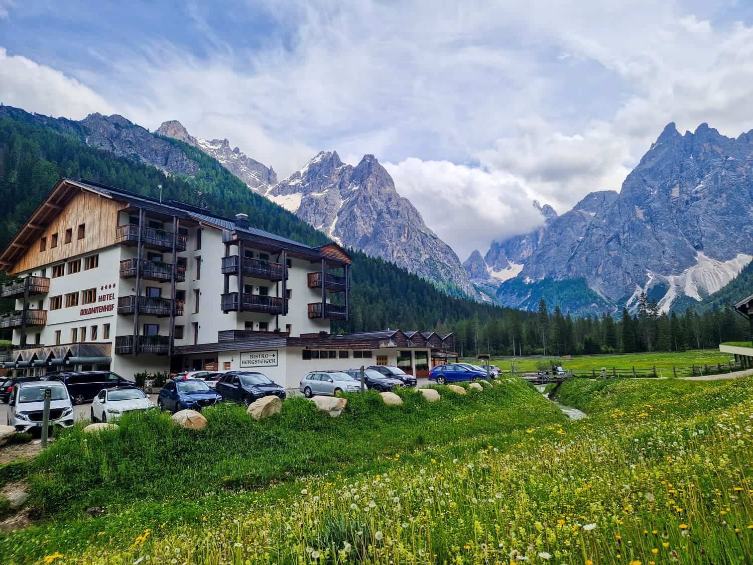 Mountain hotel in a scenic alpine valley surrounded by lush greenery and wildflowers. Cars are parked outside the multi-story wooden building. Majestic, cloud-capped mountains rise in the background under a partly cloudy sky.