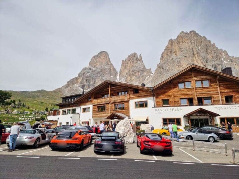 A mountain lodge named Passo Sella is situated against a rocky mountain backdrop. Several sports cars are parked in front, including red and orange vehicles. A group of people are gathered near the building under a partly cloudy sky.