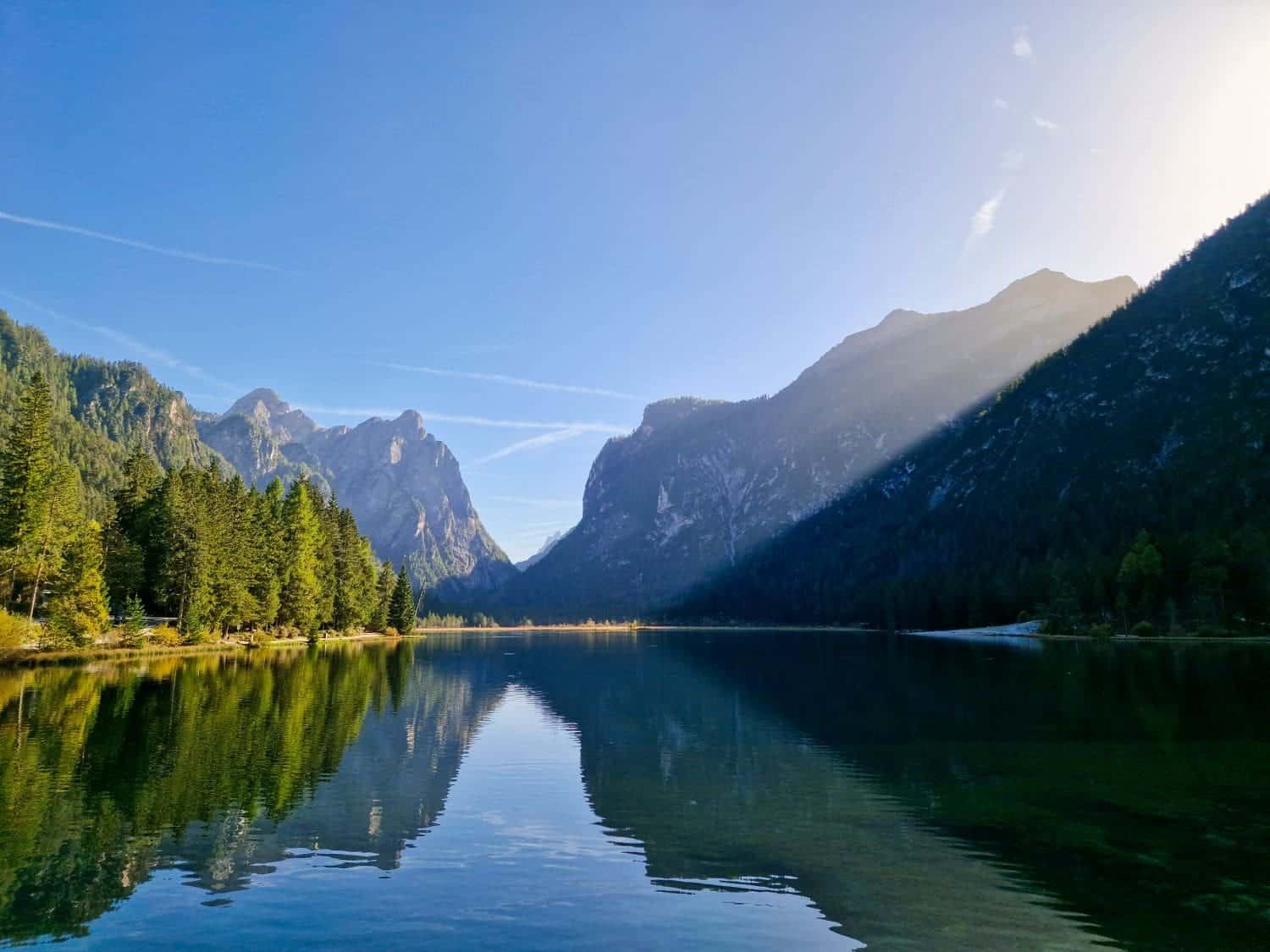 Lago di Braies: Discover The Pearl of the Dolomites 13 A serene lake reflecting surrounding pine forest and steep mountains under a clear blue sky. Sunlight casts a shadow on part of the mountain, creating a tranquil and picturesque landscape.