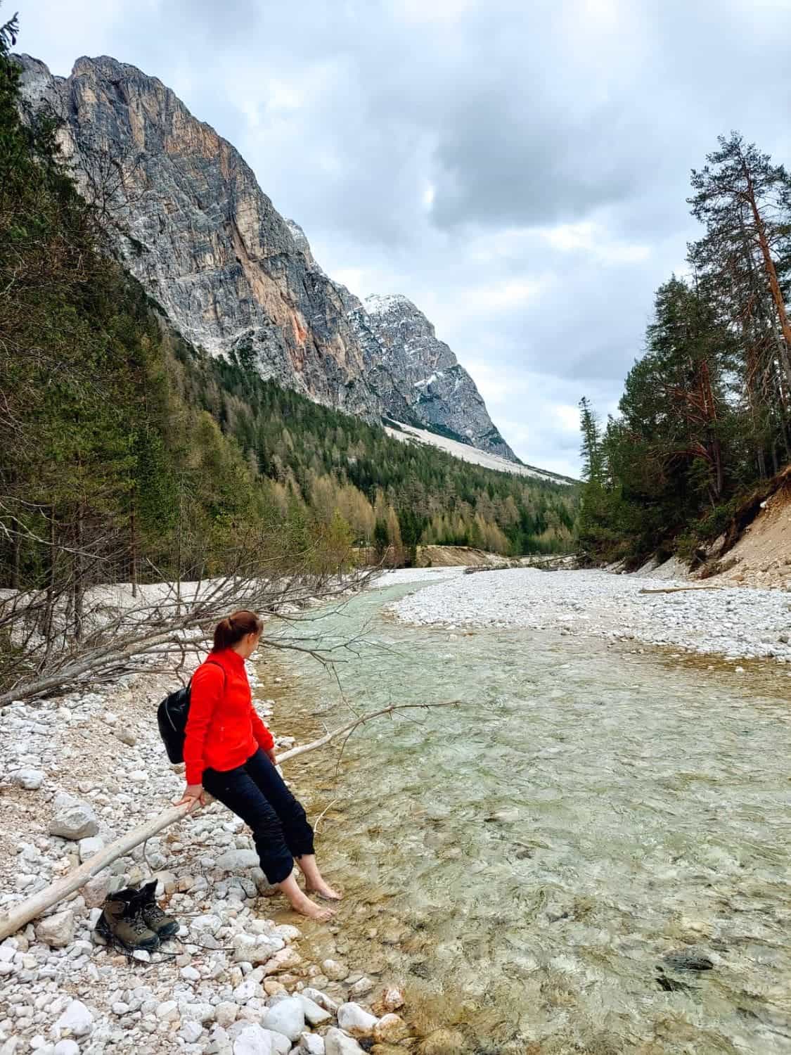 A person in a red jacket and black pants sits on a log by a clear, rocky stream, dipping their feet in the water. Pine trees and a snowy mountain are in the background under a cloudy sky.