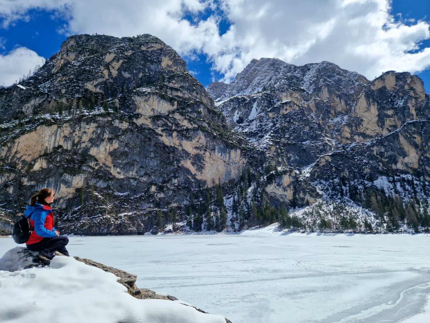 A person in a red and blue jacket sits on a rock, overlooking a frozen, snow-covered lake. Majestic, snow-dusted mountains rise in the background under a partly cloudy sky.