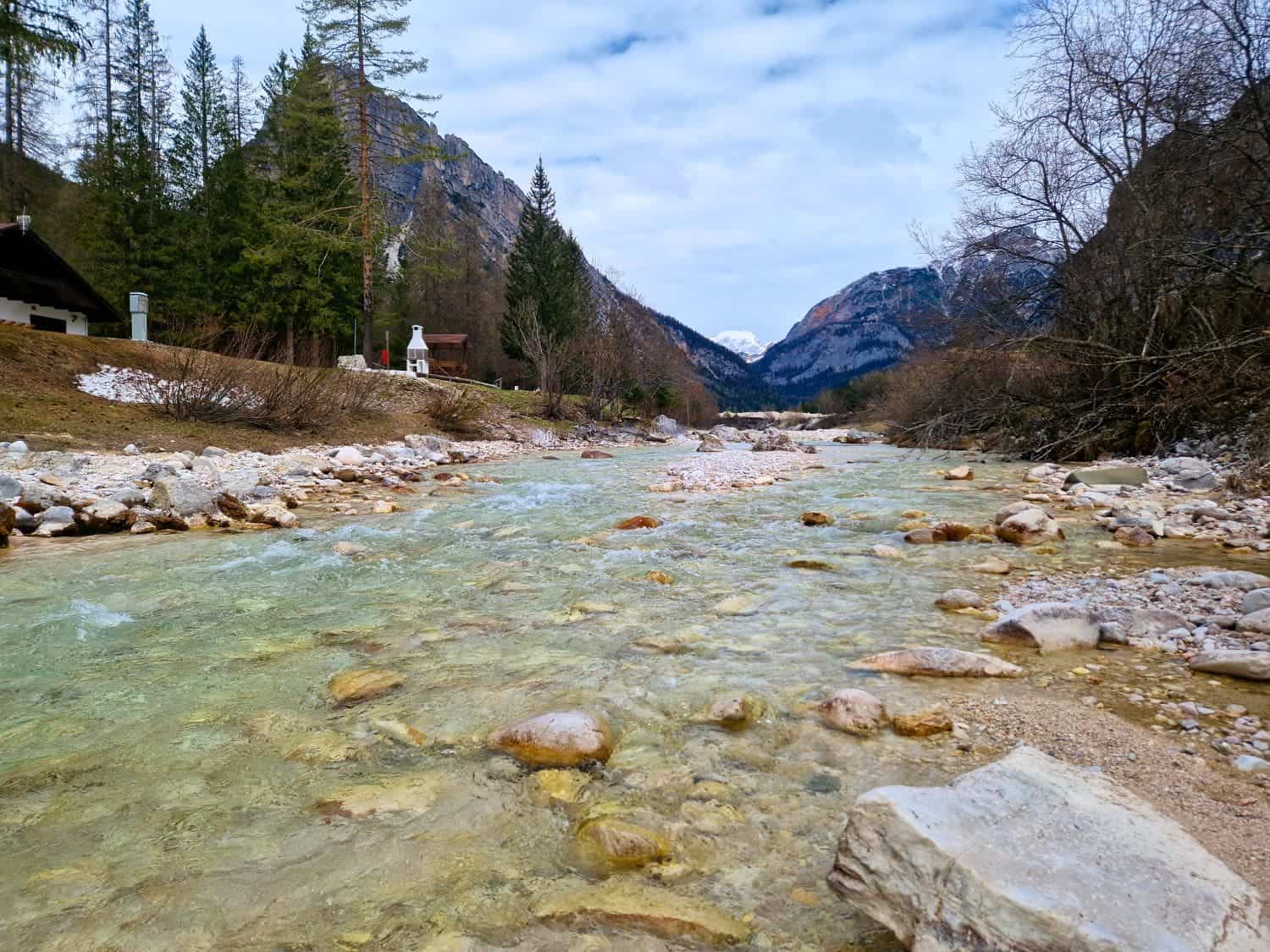 A clear, shallow stream flows through a rocky riverbed, surrounded by leafless trees and evergreen forests. Snow-capped mountains rise in the background under a cloudy sky, creating a serene, natural landscape.