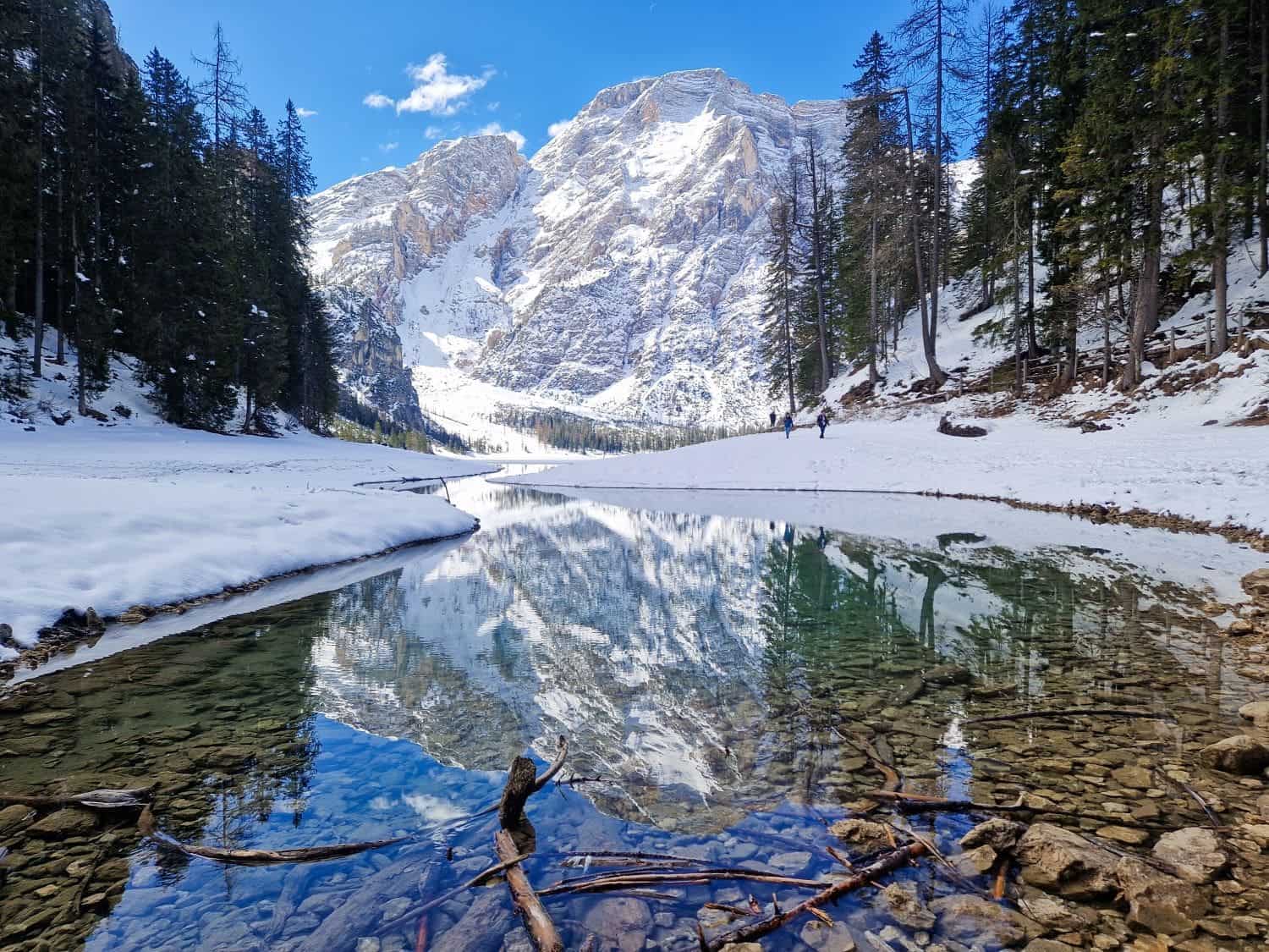 Lago di Braies: Discover The Pearl of the Dolomites 4 A clear mountain lake reflects snow-covered peaks and surrounding evergreen trees. The foreground shows stones and branches in shallow water. A few people are walking along the snowy path by the lake under a bright blue sky.