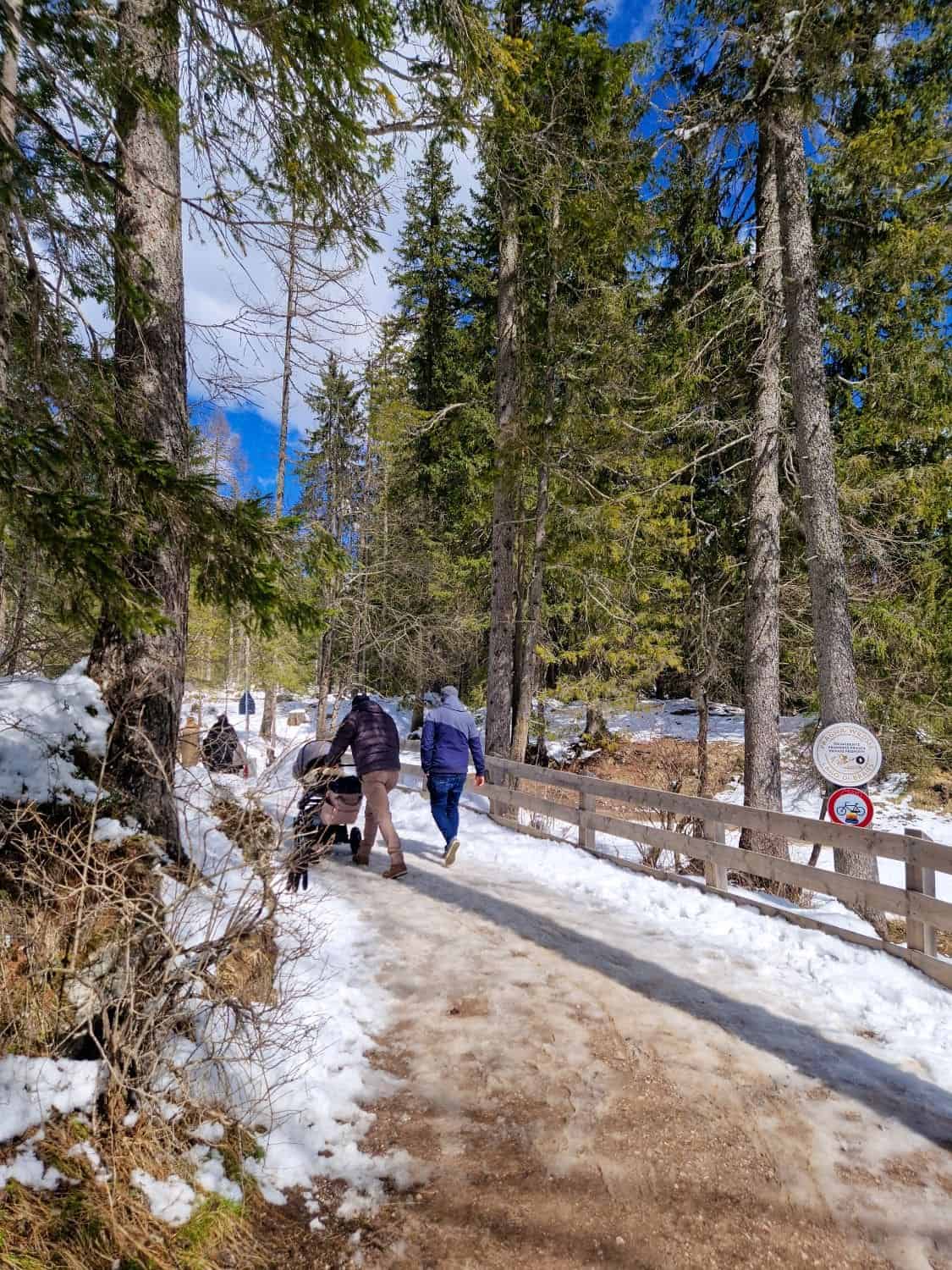 Lago di Braies: Discover The Pearl of the Dolomites 10 People walking on a snowy path through a forest with tall trees on a sunny day. Some snow covers the ground and branches. A wooden fence lines the path, and a sign is visible. The sky is blue with a few clouds.