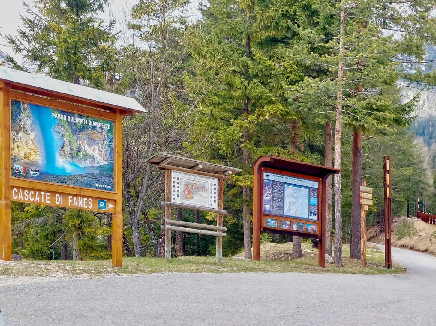 Signs and information boards at the entrance of a wooded area, including a large map labeled "Cascate di Fanes" on the left. Tall pine trees and a walking path are visible in the background.