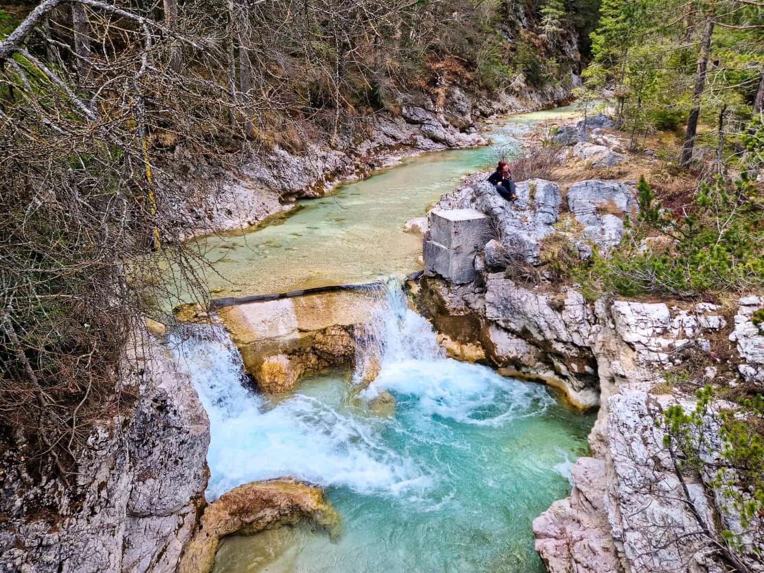 A person sits on a rock ledge near a cascading turquoise stream surrounded by rugged rock formations and dense forest. The water flows energetically, creating small rapids as it winds through the serene natural landscape.