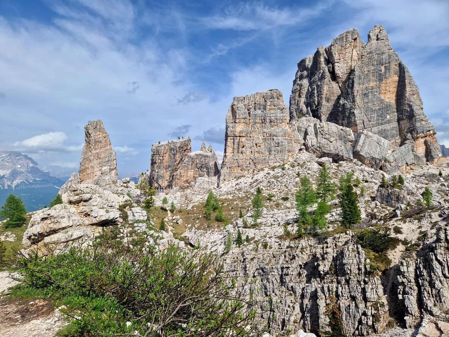 Rocky towers and rugged formations rise sharply against a partly cloudy blue sky in a mountainous terrain. Sparse greenery, including small trees and shrubs, dot the rocky landscape, creating a striking contrast with the stone.
