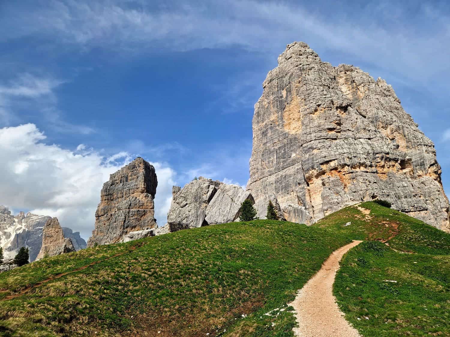A scenic view of towering rock formations in a mountainous landscape under a blue sky with clouds. A winding dirt path leads through green grass towards the rocks, with a few scattered trees along the way.
