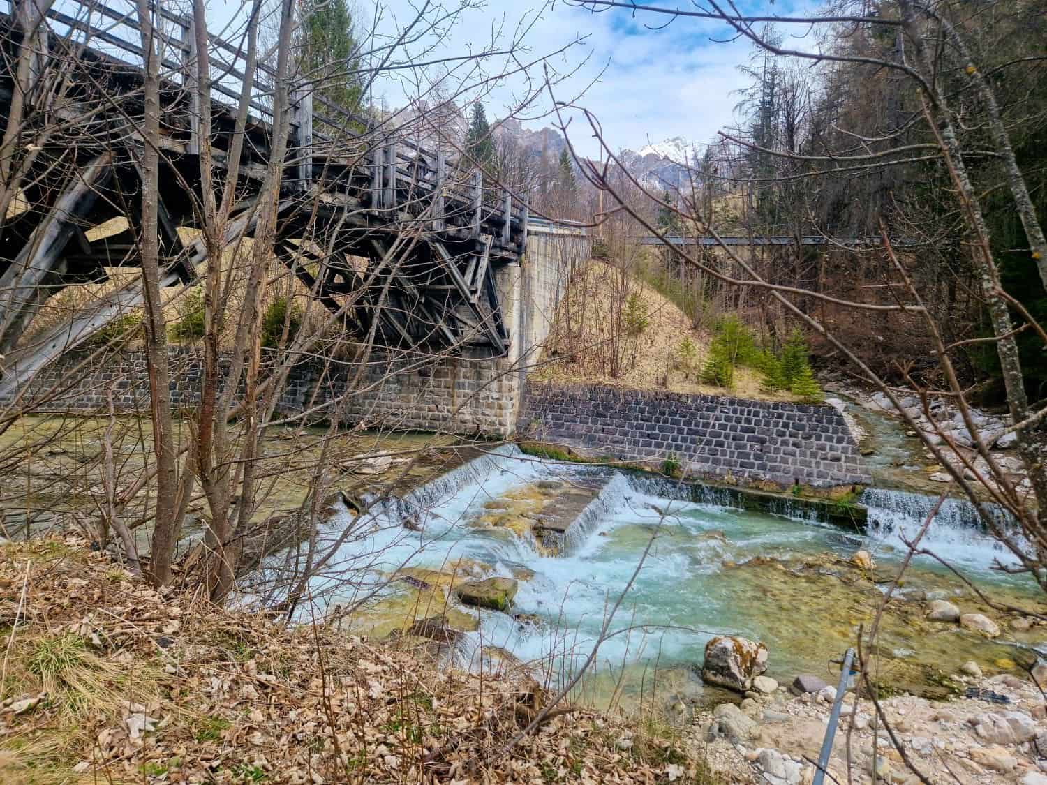 A rustic wooden bridge stretches over a rocky stream with small waterfalls. Bare trees surround the scene, and distant mountains rise against a cloudy sky. Fallen leaves cover the ground, suggesting late autumn or early spring.