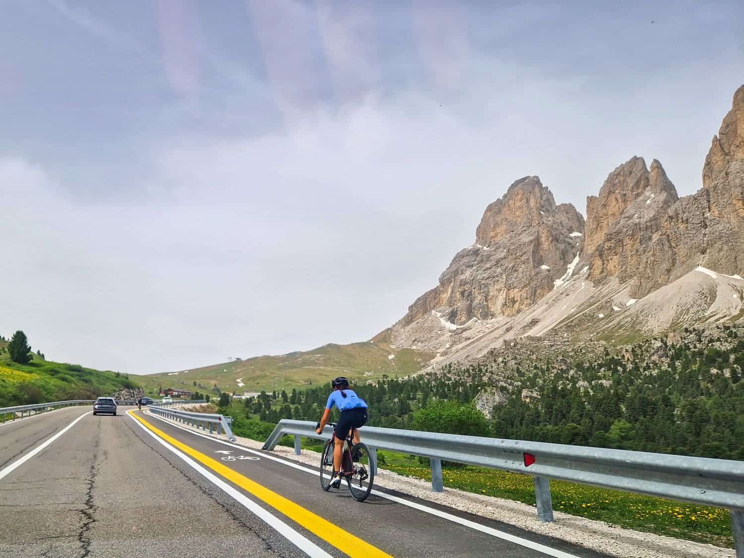 A cyclist wearing a blue shirt rides along a road surrounded by mountainous terrain. The road features distinct bike and car lanes. In the background, large rocky peaks rise under a partly cloudy sky.