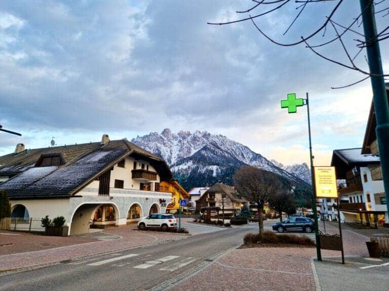A picturesque street scene with snow-capped mountains in the background. Traditional Alpine houses line the road, and a pharmacy with a green cross sign is visible. A car is parked, and the sky is partly cloudy. Trees and a pedestrian crossing are present.
