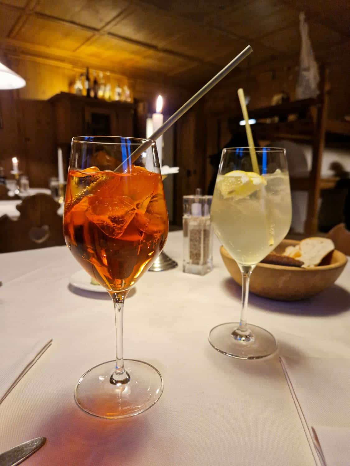 Two glasses of cocktails on a table with a white tablecloth. One glass contains an orange drink with ice and an orange slice; the other a light-colored drink with ice and a lemon slice. A bowl of bread is in the background, with a cozy, dimly-lit setting.
