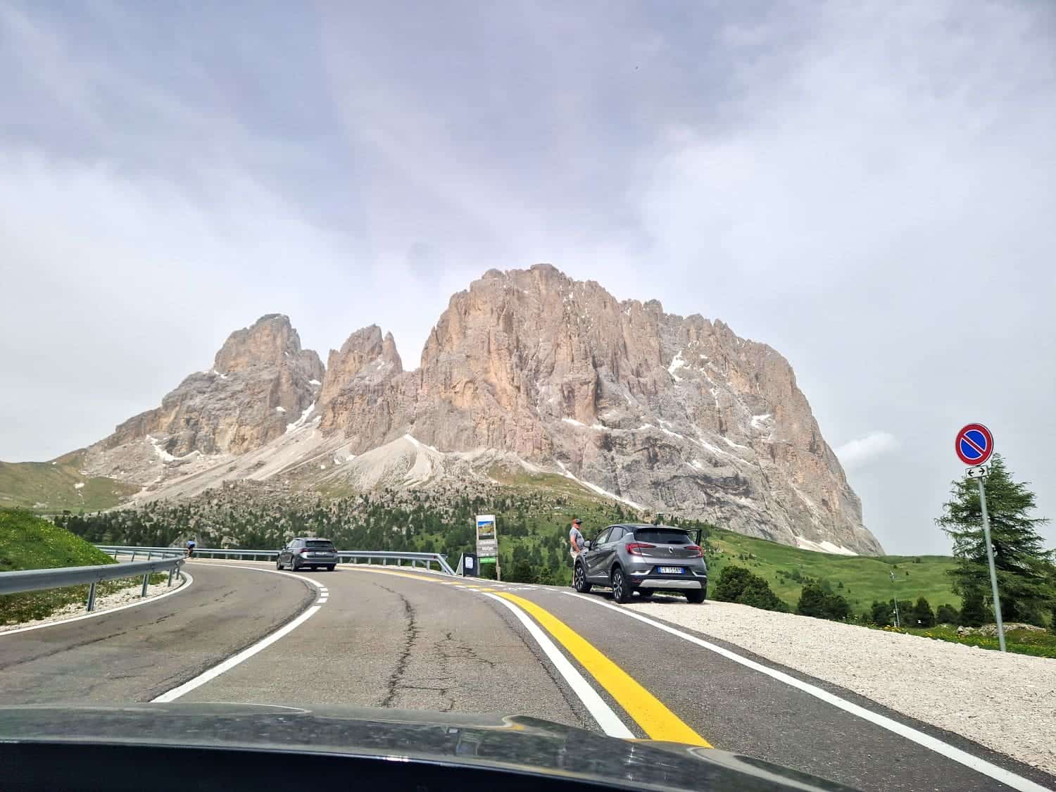 A winding road leads to majestic, rocky mountains under a cloudy sky. Cars are parked along the roadside, with one person visible near a parked car. Lush green grass contrasts the rugged mountain terrain. A no-parking sign is also visible.