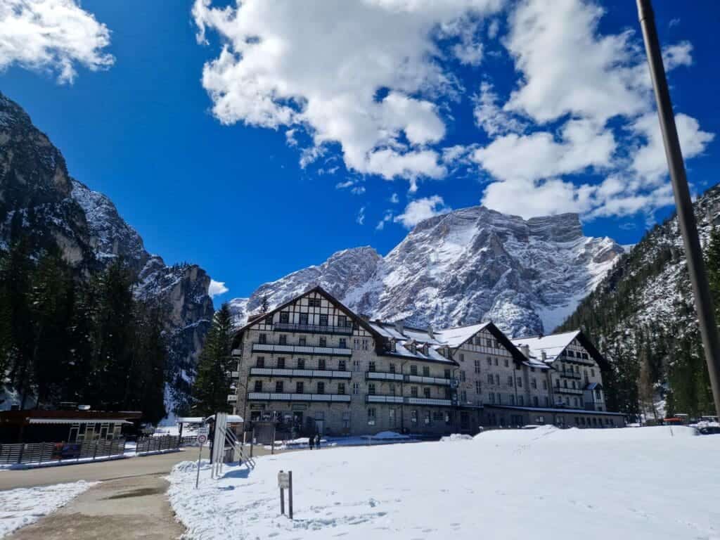 A large, alpine-style hotel stands amidst a snowy landscape with towering mountains in the background. The sky is bright blue with scattered fluffy clouds. Pine trees flank the building, adding to the picturesque, serene setting.