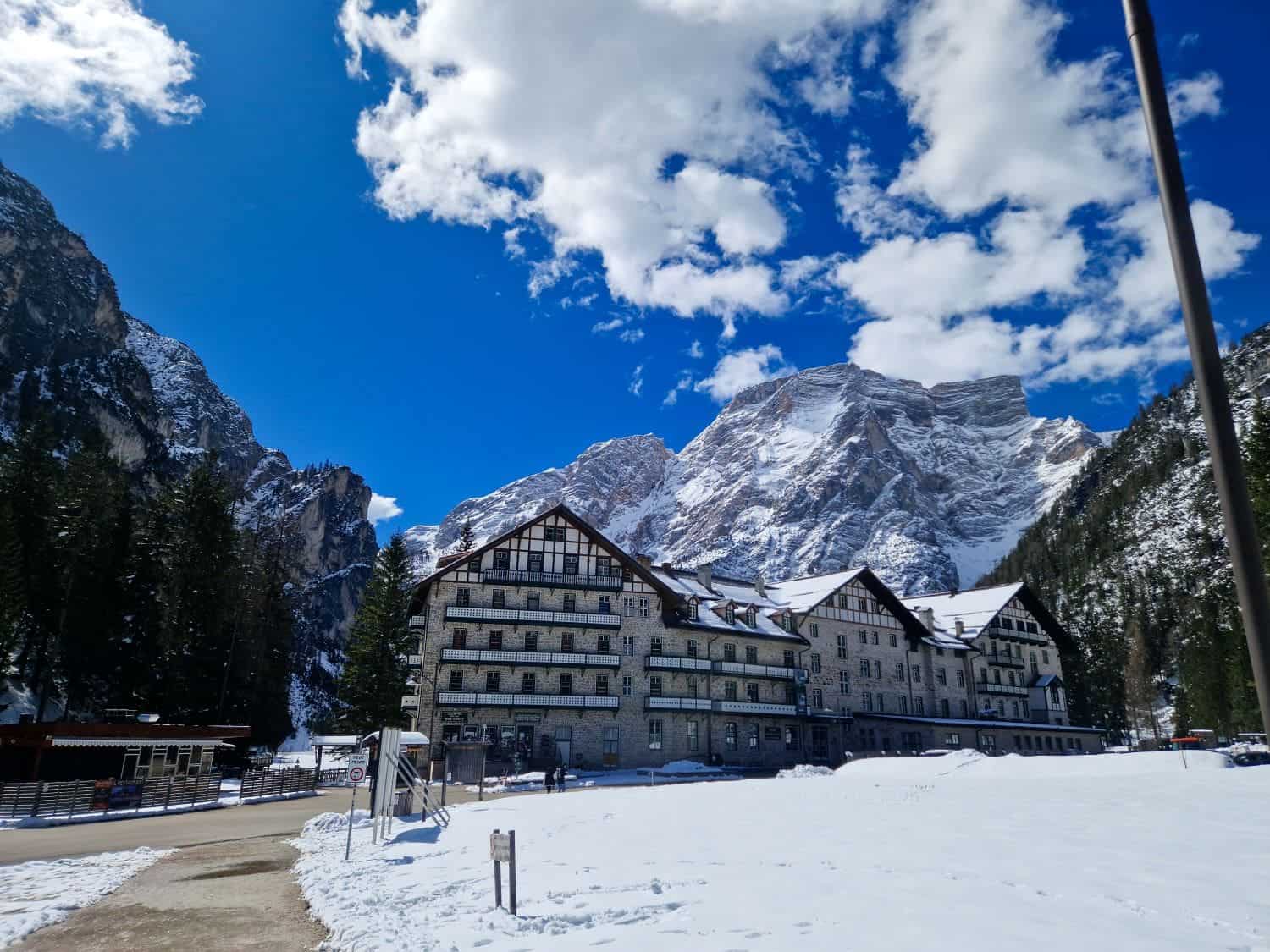 Lago di Braies: Discover The Pearl of the Dolomites 14 A large, alpine-style hotel stands amidst a snowy landscape with towering mountains in the background. The sky is bright blue with scattered fluffy clouds. Pine trees flank the building, adding to the picturesque, serene setting.