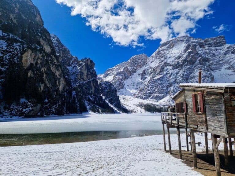 Snow-covered mountains and a wooden cabin beside a frozen lake under a bright blue sky with scattered clouds. The scene is serene, with a mix of sunlight and shadows highlighting the rugged landscape.
