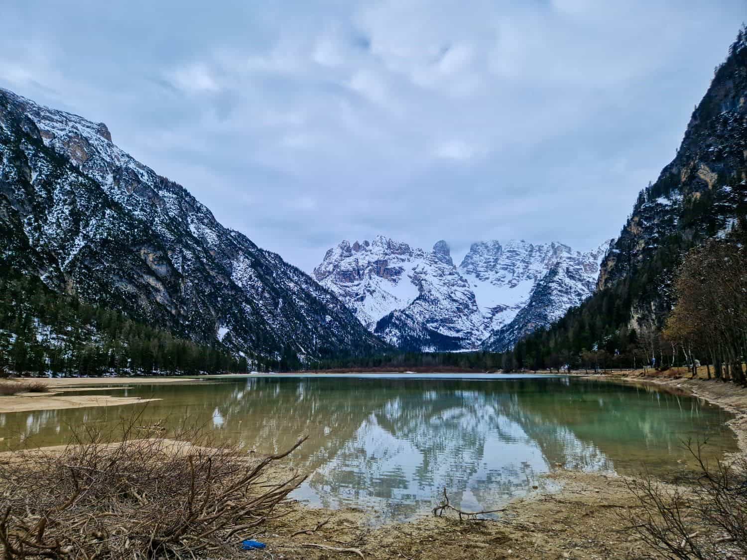 A serene mountain lake with clear reflections of snow-capped peaks and surrounding forests under a cloudy sky. The landscape is framed by rugged mountains on both sides, with a mix of bare and evergreen trees lining the shore.