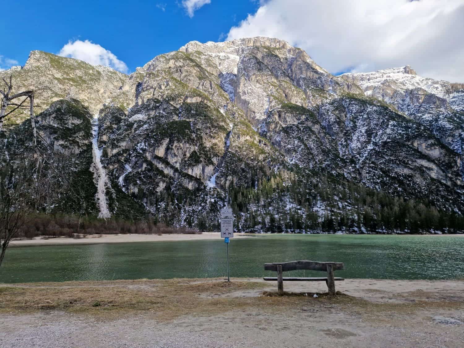 A serene mountain landscape featuring a calm lake in the foreground and snow-capped peaks under a blue sky with clouds. A wooden bench faces the lake, and a signpost stands nearby, adding a human element to the natural scene.