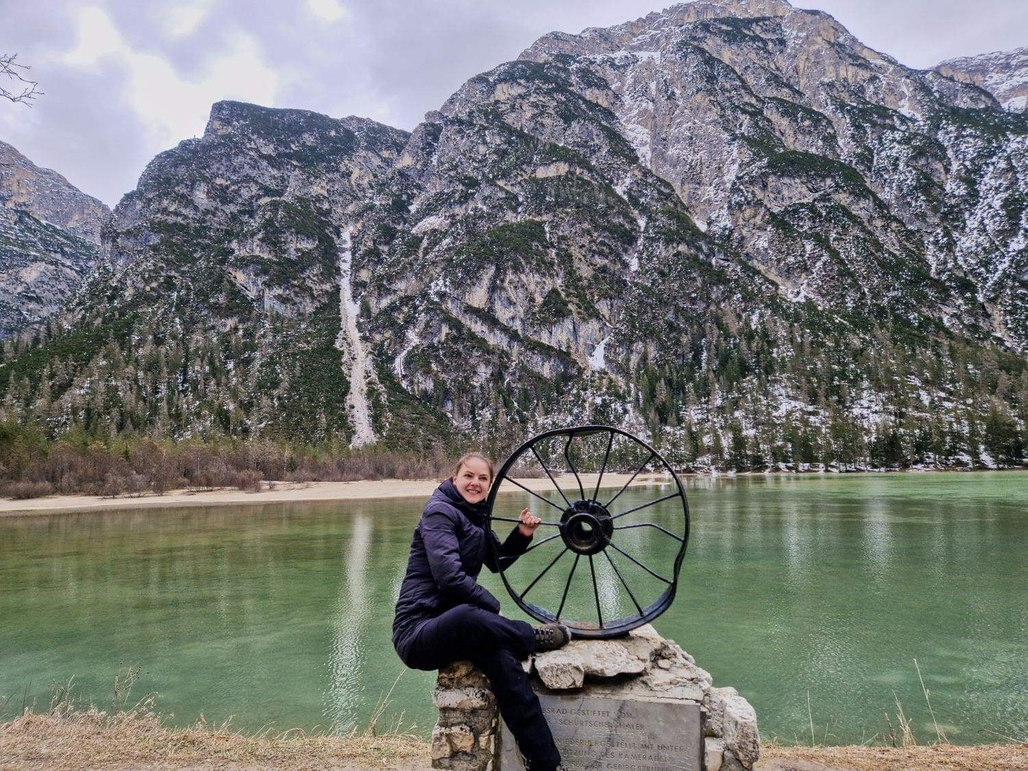 A person in a black jacket sits on a rock near a scenic lake, holding a large wheel sculpture. Snow-dusted mountains and evergreen trees frame the background, under a cloudy sky.