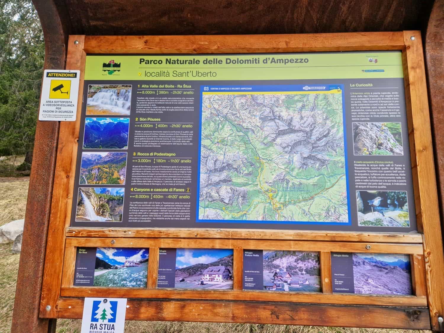 Information board at Parco Naturale delle Dolomiti d'Ampezzo, locality Sant’Uberto. It features maps, hiking routes, and local regulations. Below are smaller images of park views. The sign is wooden with a natural background.