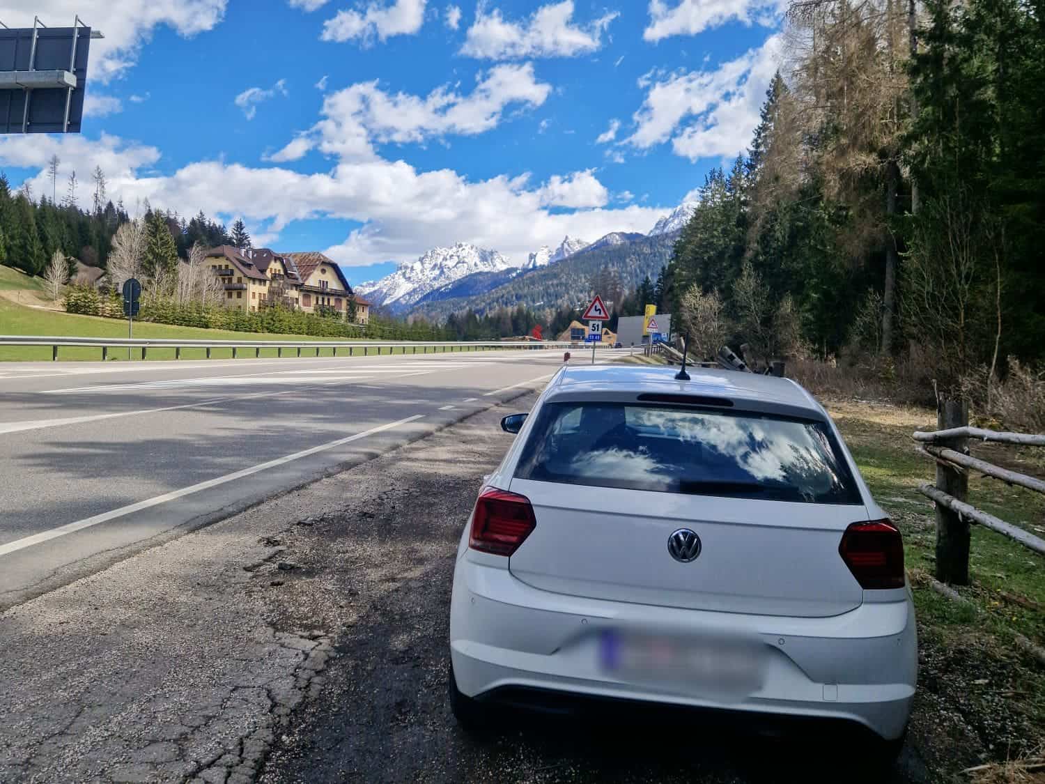 A white car is parked on the side of a scenic road under a blue sky with scattered clouds. Snow-capped mountains and evergreen trees line the background. A few buildings are visible on the left.