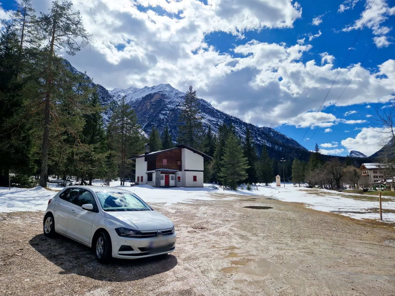 A white car is parked on a dirt path near a house surrounded by snow. Tall pine trees and a mountain range are in the background under a partly cloudy sky.