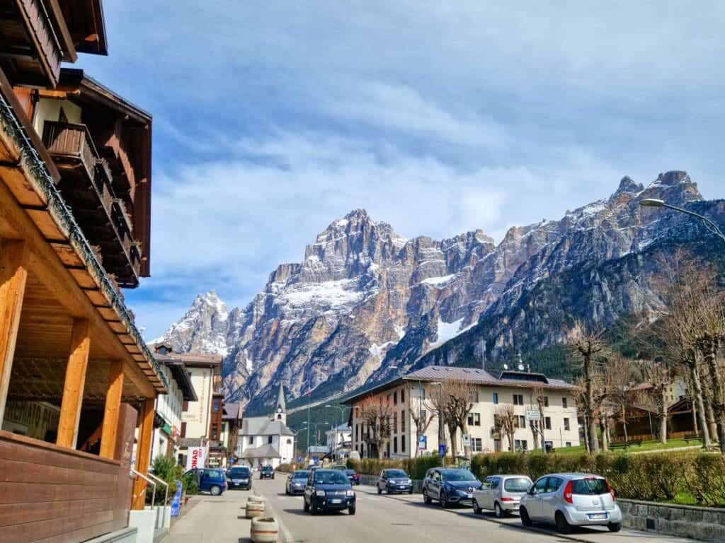 A picturesque street in a mountain town with wooden houses and buildings. Snow-capped peaks are visible in the background under a partly cloudy sky. Cars are parked along the street, and the scene has a peaceful, vibrant atmosphere.