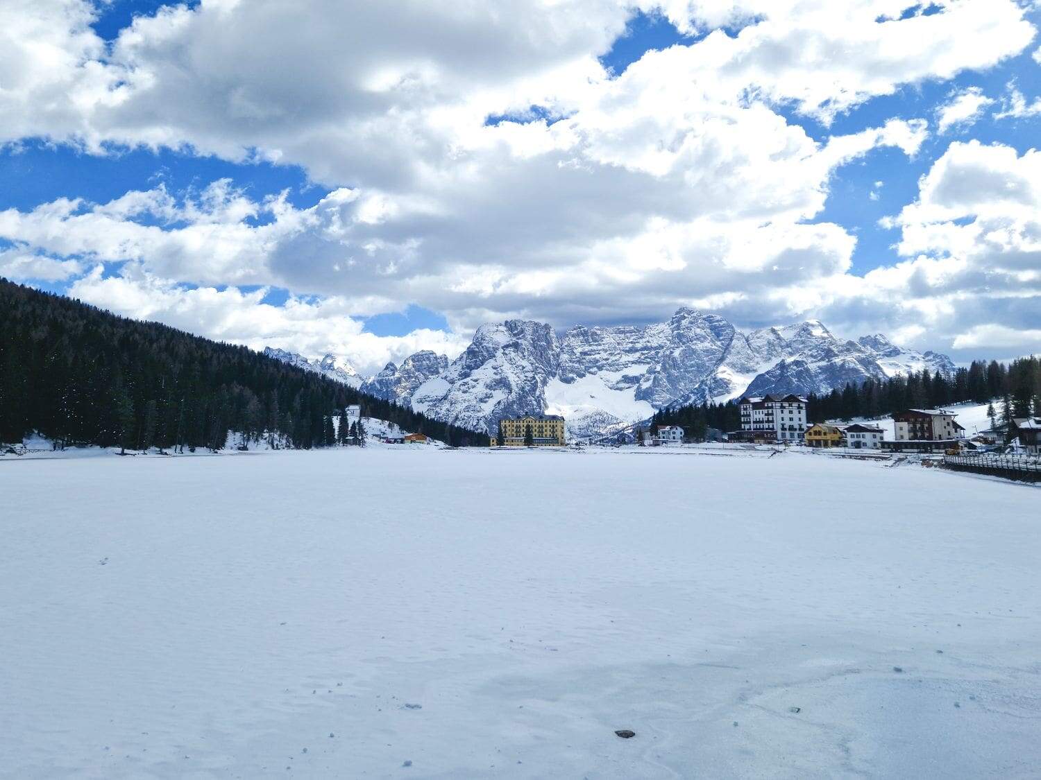 A snowy landscape featuring a frozen lake in the foreground, with buildings nestled among trees on the right. towering snow-covered mountains in the background under a partly cloudy blue sky.