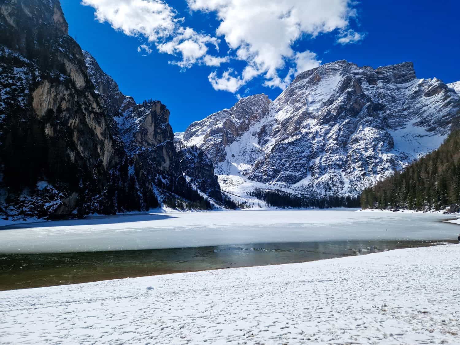 Snow-covered mountains surround a partially frozen lake under a vibrant blue sky with scattered clouds. A thin layer of snow covers the ground in the foreground. Pine trees line the right side of the landscape.