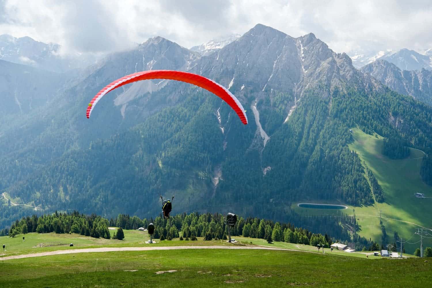 A person paraglides with a red paraglider over a lush green meadow, surrounded by tall, rugged mountains under a partly cloudy sky. The scene captures a sense of adventure and natural beauty.
