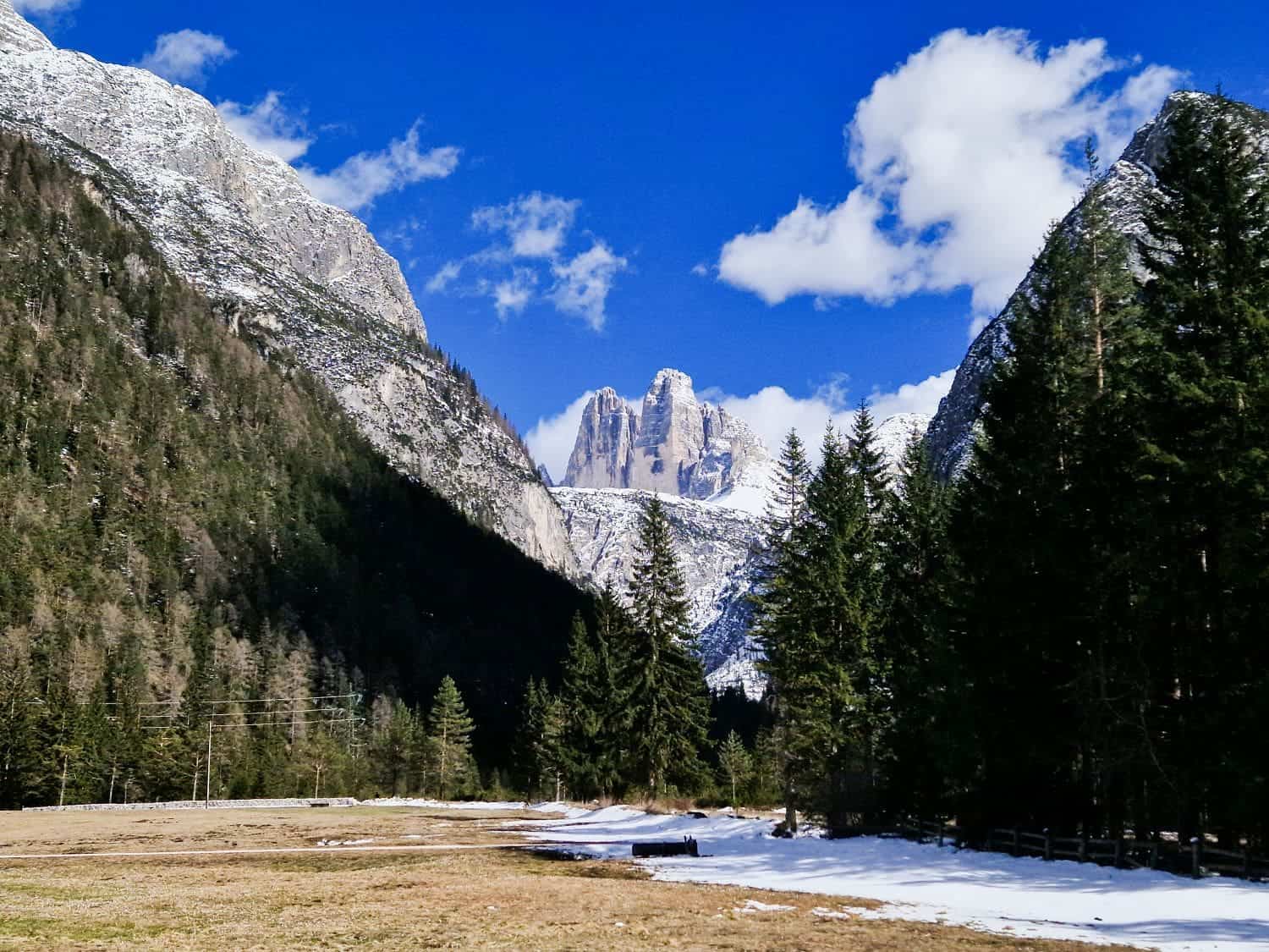 A scenic view of a mountainous landscape with snow-capped peaks under a blue sky. Pine trees frame the foreground, with patches of snow on the ground. Fluffy clouds float above, adding contrast to the vibrant scene.