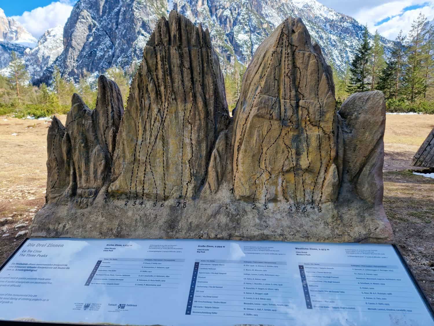A rock display resembling a mountain range with numbered climbing routes is set against a backdrop of real mountains. A sign in front provides information about the climbing paths. Snow-capped peaks and trees are visible in the background.
