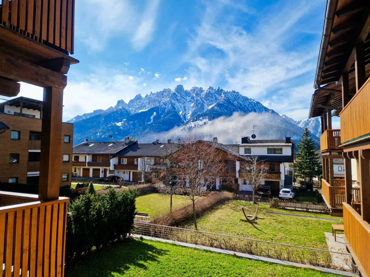 Mountain view from a wooden balcony with residential houses in front. Snow-capped peaks under a partly cloudy sky create a scenic backdrop. A grassy yard with a swing is visible between the buildings.