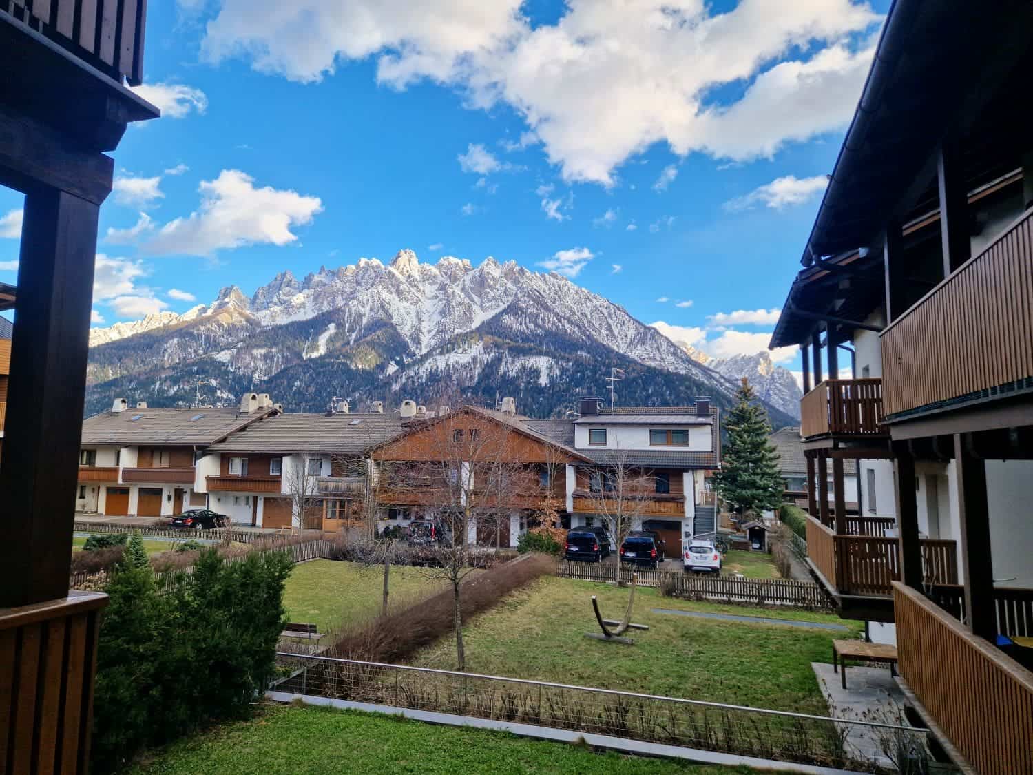 View of a snowy mountain range under a partly cloudy sky, framed by wooden residential buildings. A grassy yard with bare trees is visible, along with a few parked cars in front of the houses.