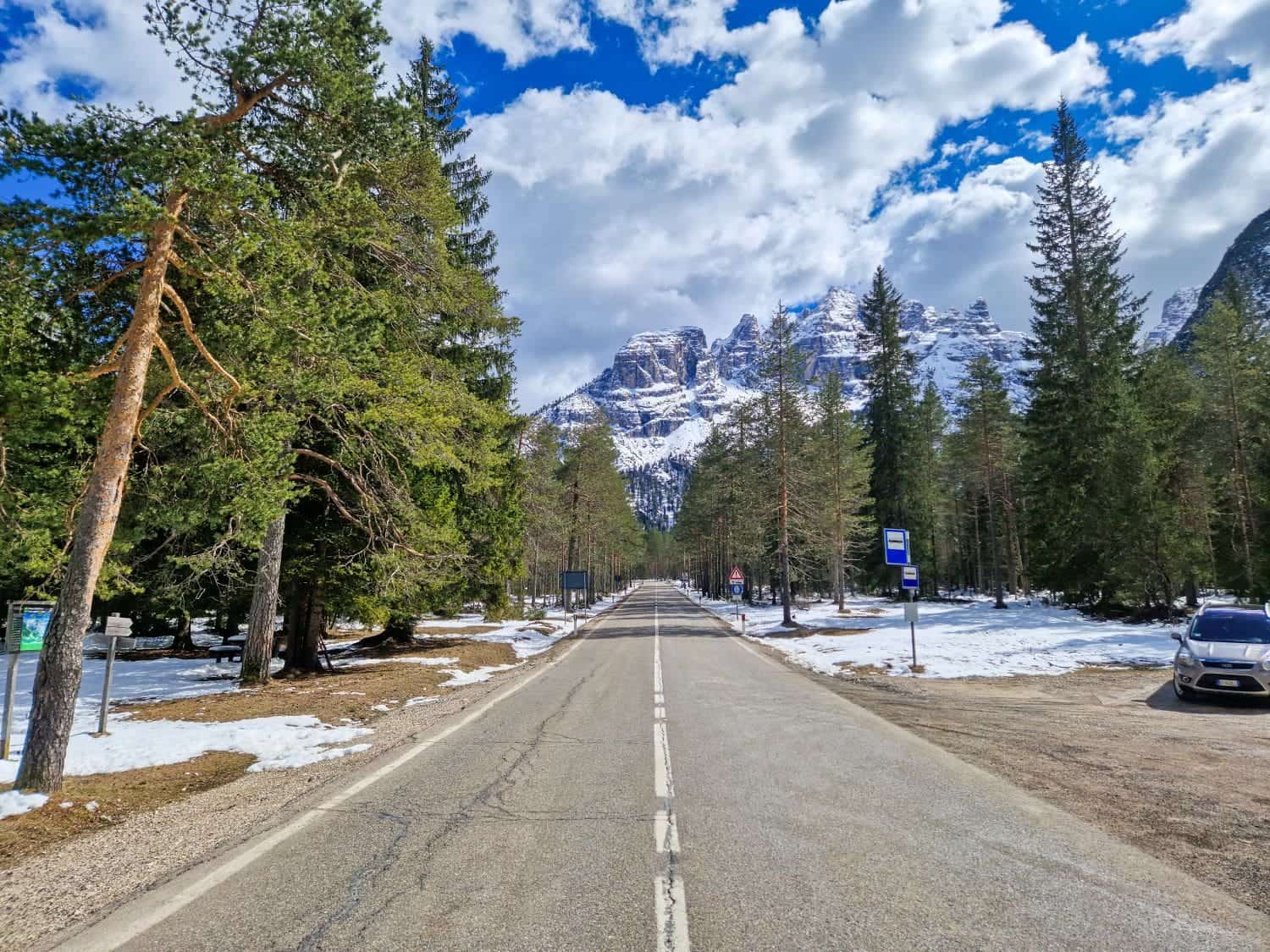A straight road in a snowy forest leads towards distant snow-covered mountains under a partly cloudy blue sky. Trees line the roadside, and a car is parked in a small lot to the right. Signs and patches of melting snow are visible.