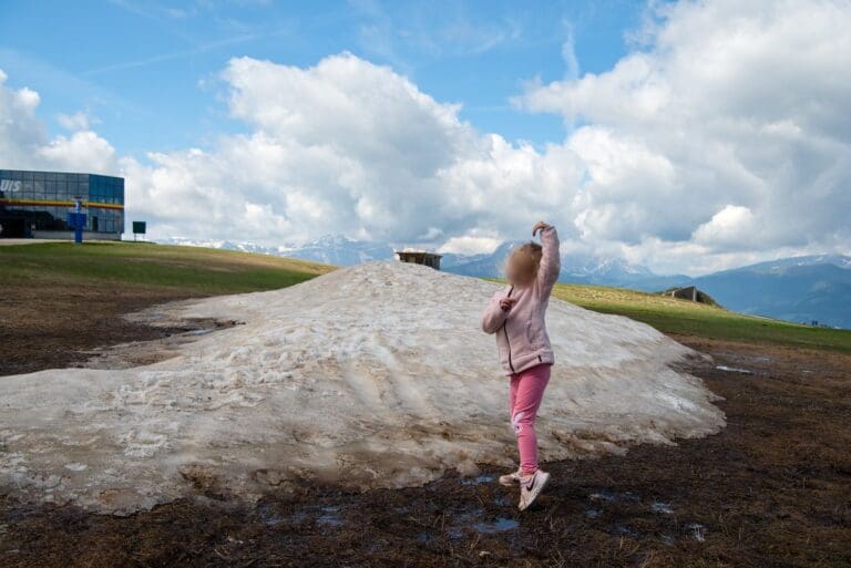 A child in a pink outfit stands on a patch of melting snow on a grassy field. Mountains and a cloudy sky are in the background, with a building visible on the left.