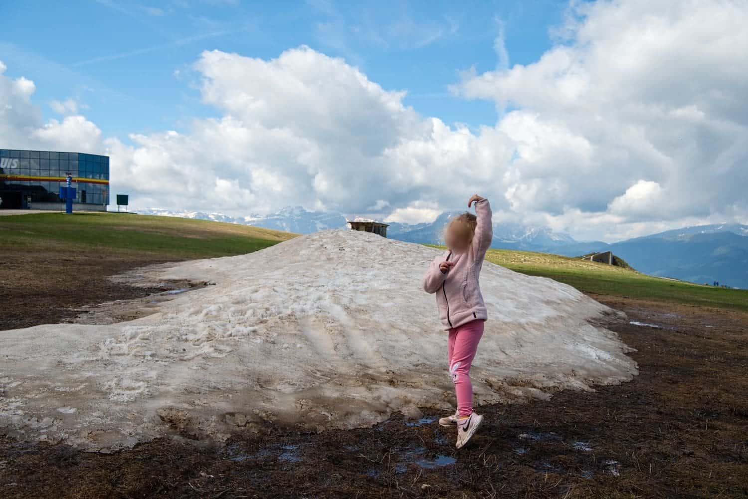 A child in a pink outfit stands on a patch of melting snow on a grassy field. Mountains and a cloudy sky are in the background, with a building visible on the left.