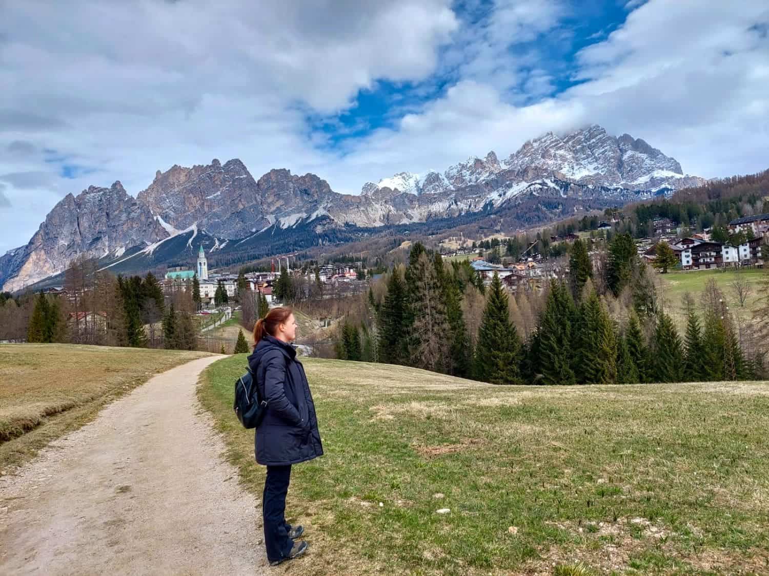 Lago di Sorapis Hike: Most Incredible Lake In The Dolomites 14 A person in a dark coat and backpack stands on a dirt path, gazing at a mountainous landscape dotted with trees and houses. Snow-capped peaks rise in the distance under a partly cloudy sky.