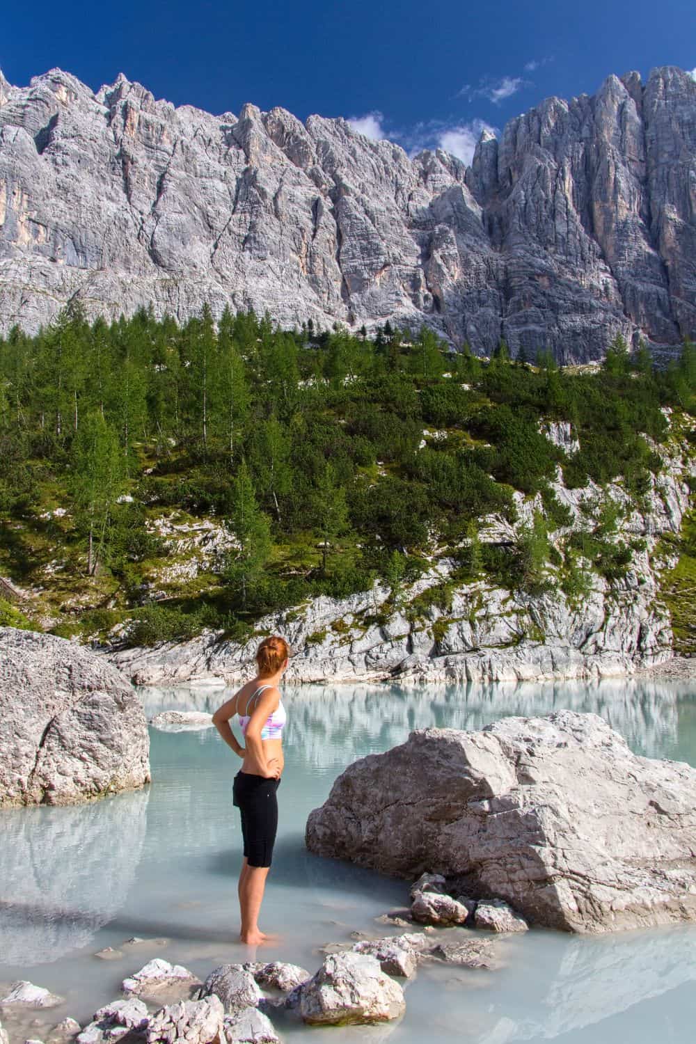 Lago di Sorapis Hike: Most Incredible Lake In The Dolomites 11 A woman stands on rocks at the edge of a serene, turquoise lake. She gazes towards towering, rugged mountains under a bright blue sky. The landscape is lush with green trees. She is wearing activewear and has her hands on her hips.