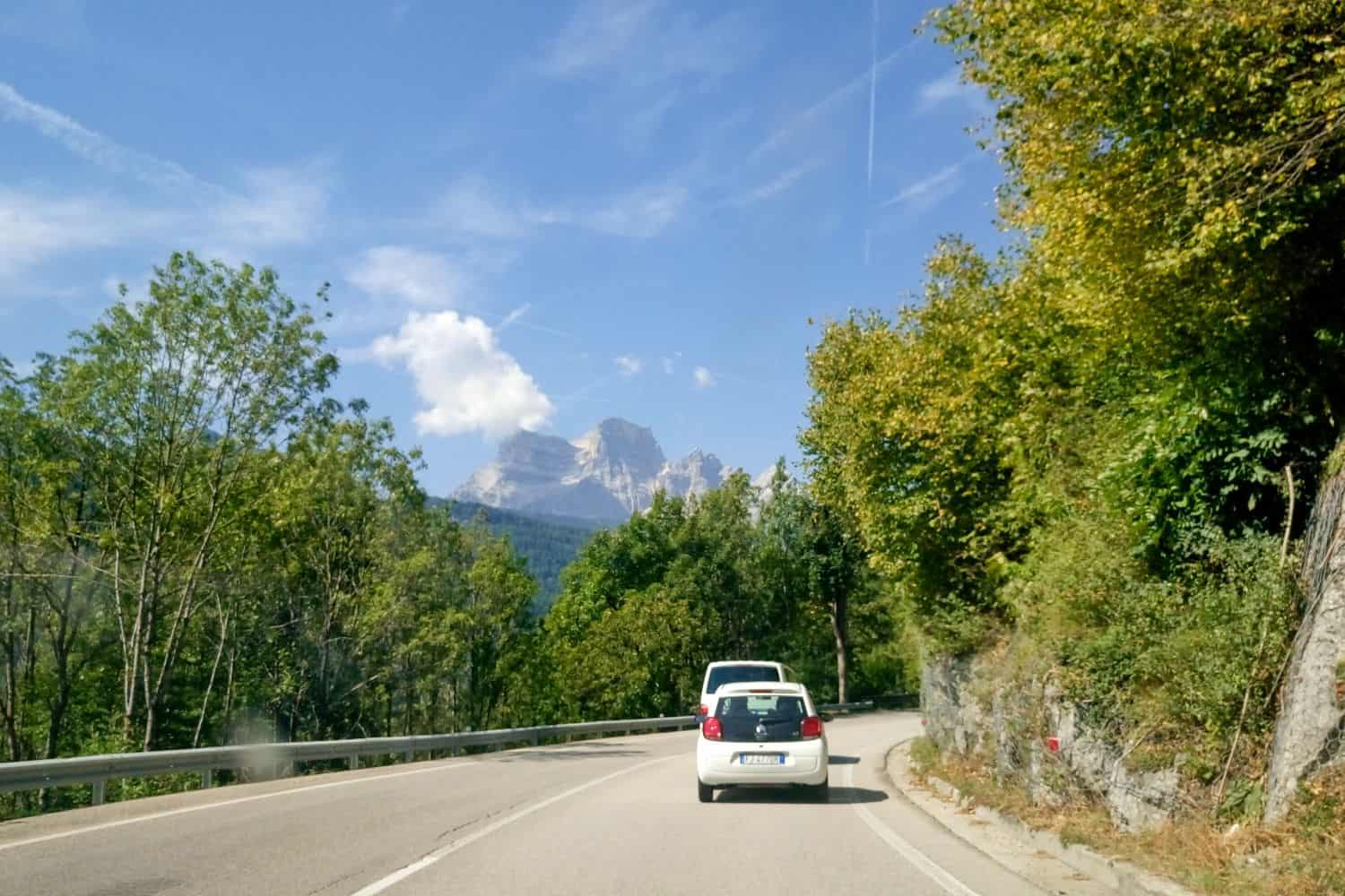 Lago di Sorapis Hike: Most Incredible Lake In The Dolomites 10 A white car drives on a winding road bordered by green trees. In the background, a mountain with a partly cloudy sky is visible. The road curves gently to the left, and the scene is under a bright, clear sky.
