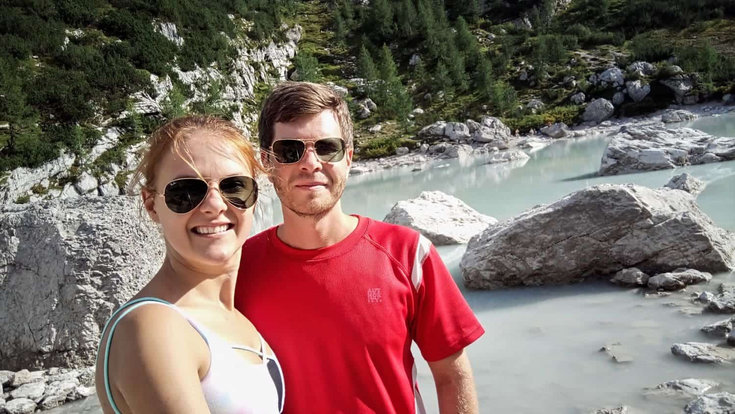 Lago di Sorapis Hike: Most Incredible Lake In The Dolomites 3 A smiling couple wearing sunglasses poses for a selfie in front of a rocky landscape with a serene, milky blue lake. Green vegetation covers the slopes in the background.