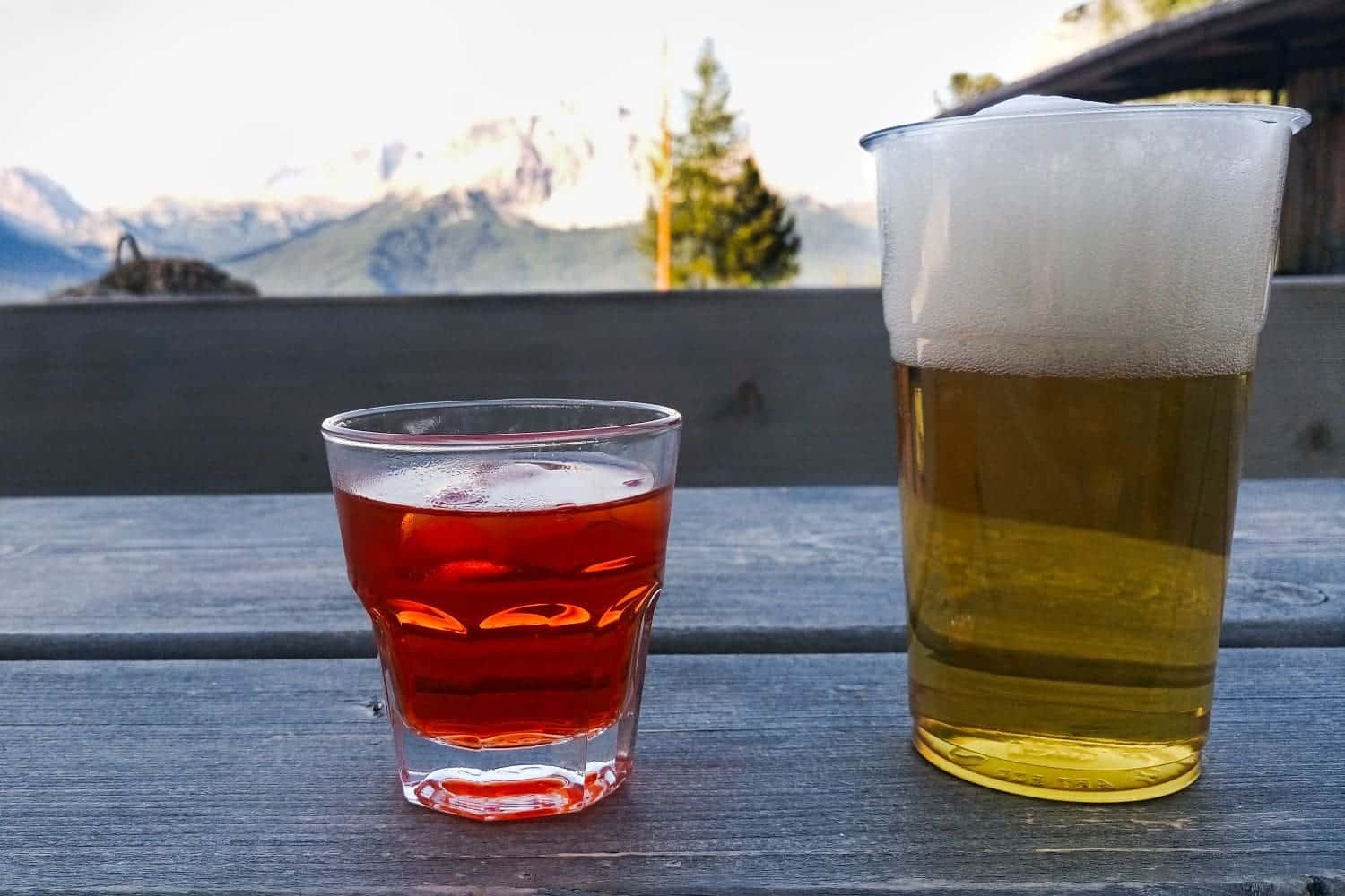 Lago di Sorapis Hike: Most Incredible Lake In The Dolomites 9 A glass of orange-colored aperitif with ice and a glass of frothy beer sit on a wooden table. In the background, there are blurry views of mountains and trees under a clear sky.
