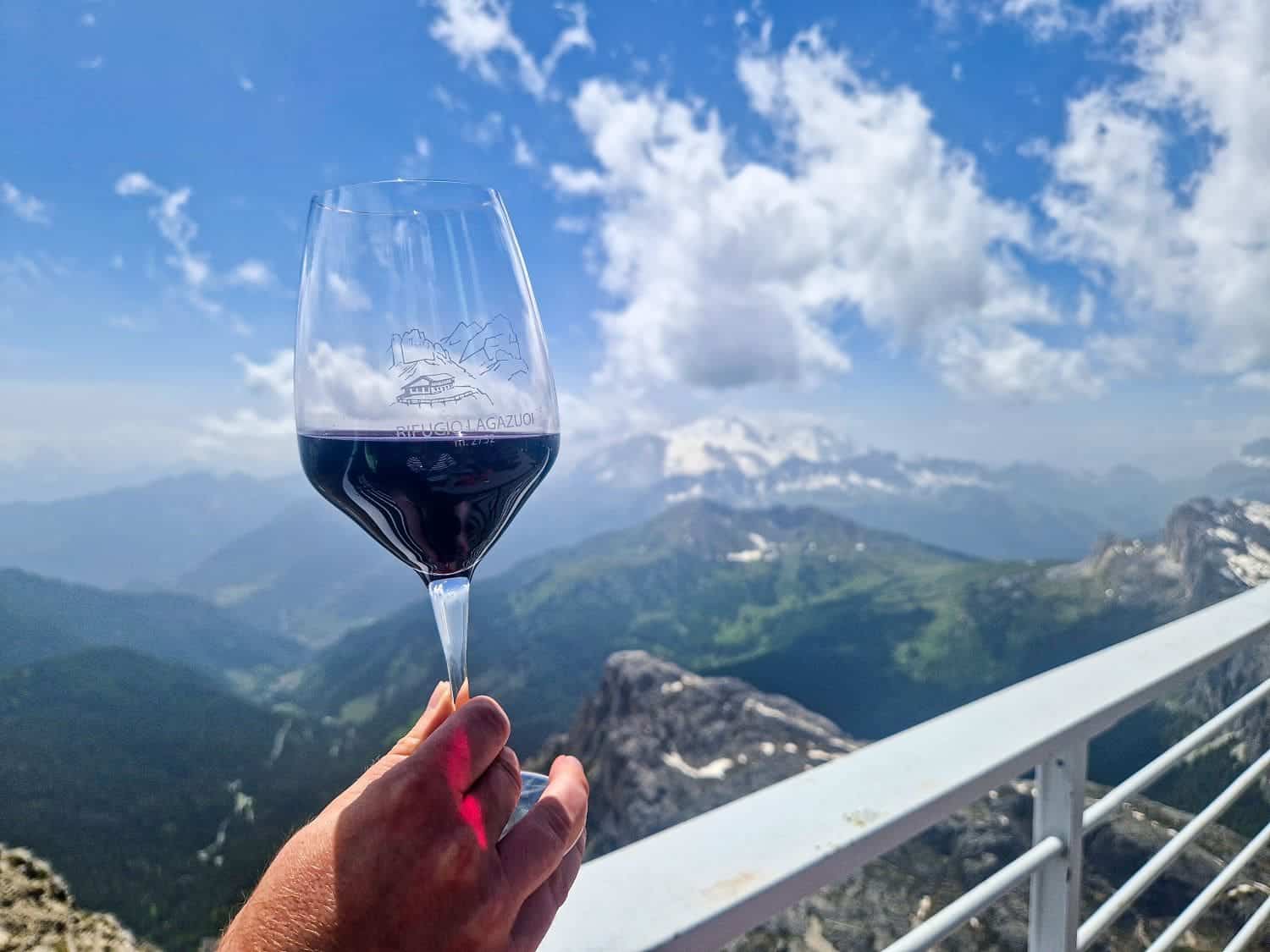 A person holds a glass of red wine against a backdrop of a mountainous landscape with lush green valleys and snow-capped peaks under a blue sky with scattered clouds. The glass features a mountain logo.