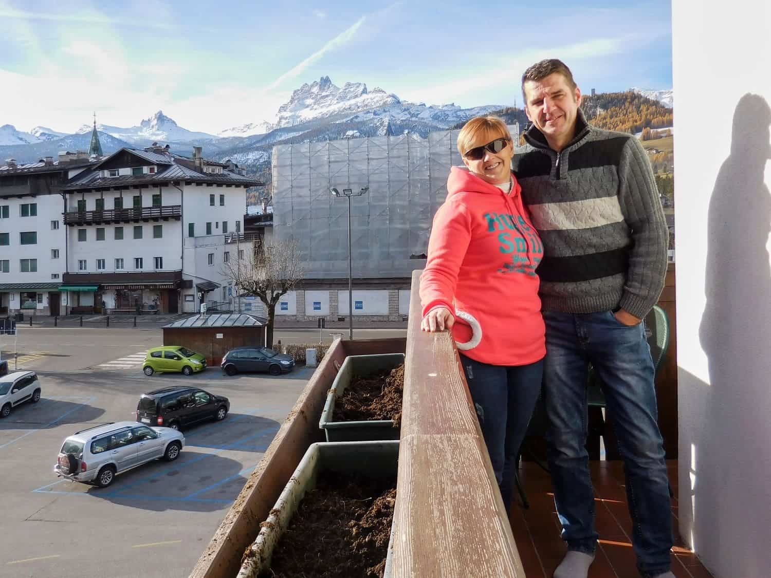 A man and woman stand on a balcony smiling. The woman wears a pink hoodie and sunglasses, and the man wears a gray sweater. Behind them, there are mountains, buildings, and parked cars under a clear blue sky.