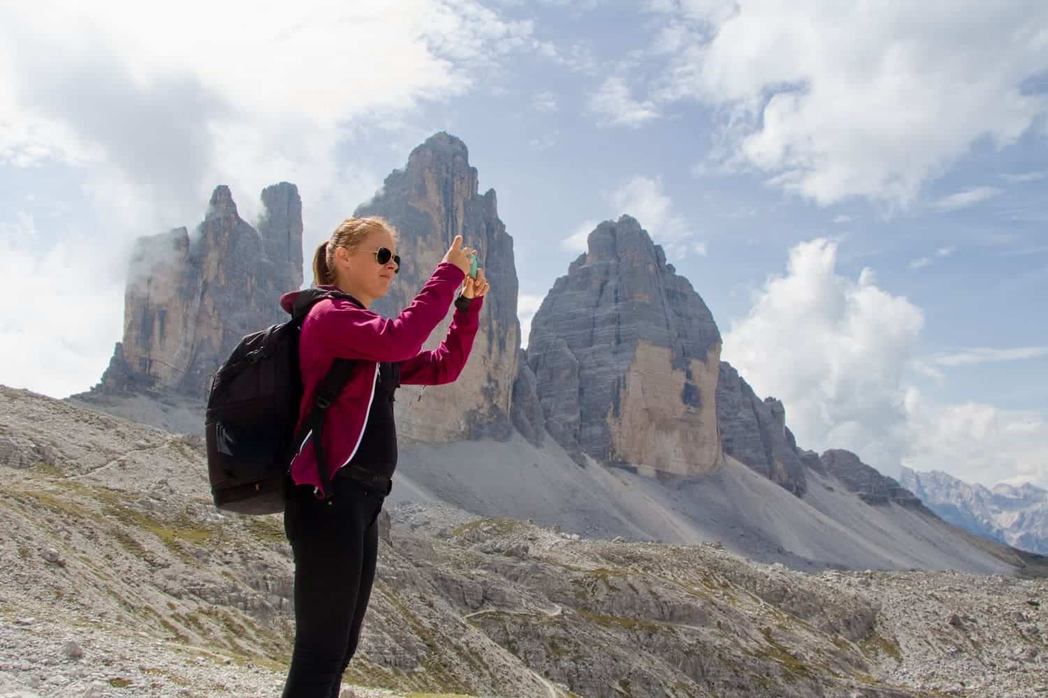 A person wearing sunglasses, a pink jacket, and a black backpack takes a photo with a smartphone. Behind them are the rocky peaks of a mountainous landscape under a partly cloudy sky.