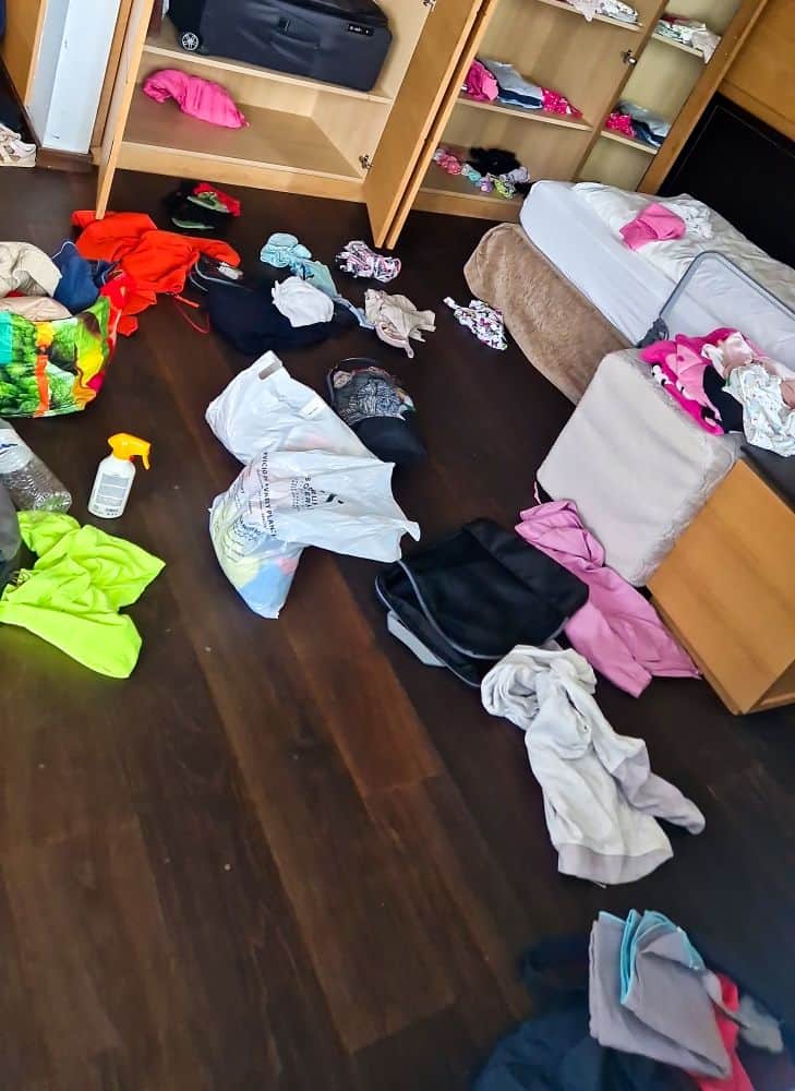A messy room with clothes scattered on a dark wooden floor. An open wardrobe holds more clothes and luggage. A bed with a beige blanket is partially visible, with bags and garments strewn around.